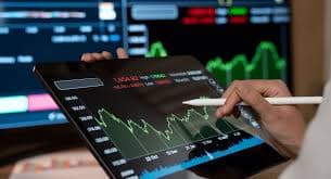Hands using a stylus on a tablet displaying financial charts with background data monitors.